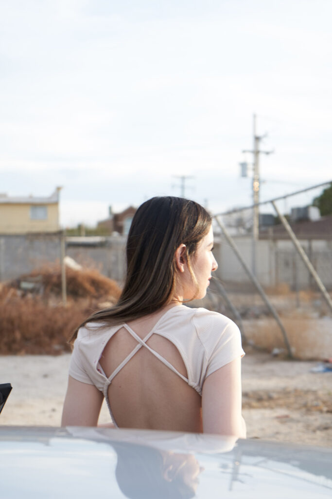 Gabriela Inzunza for Avice. A side profile of a brunette with long hair wearing a white top with a unique cut-out design gazing into the distance. The backdrop consists of a fenced area with buildings and utility poles reflecting a somewhat desolate or industrial setting.